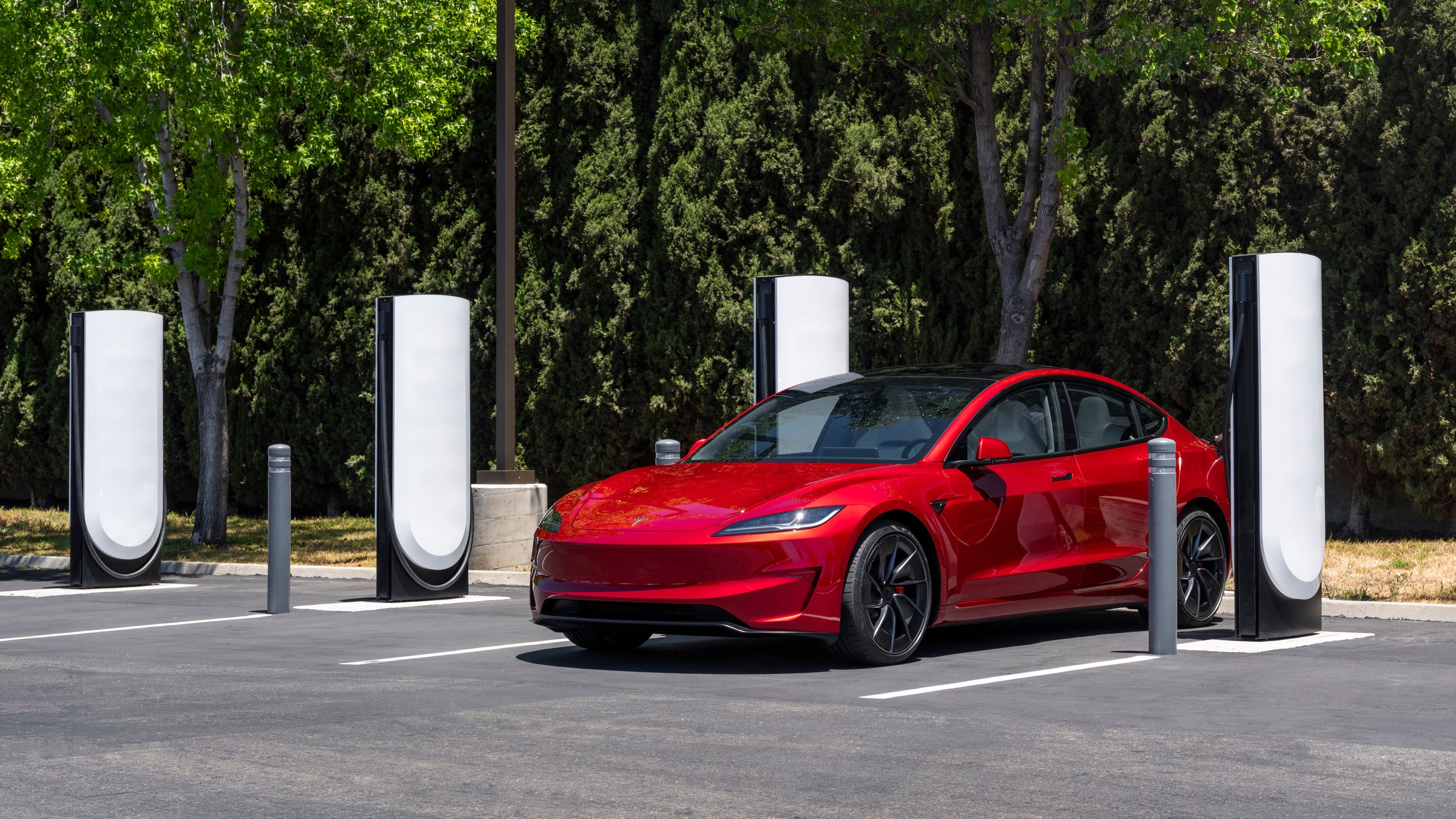 A red Tesla Model 3 parked beside V4 Supercharger stalls in a tree-lined lot