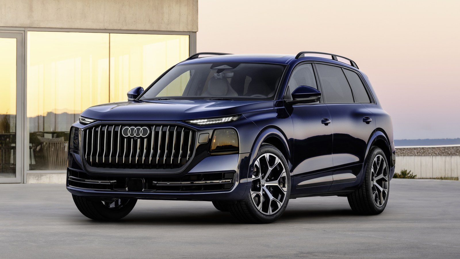 2027 Audi Q9 three-row SUV in dark blue, front three-quarter view, parked outside a modern concrete building at dusk