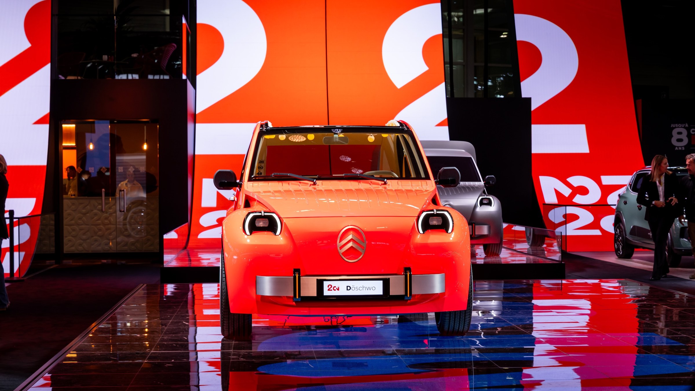Orange Citroën 2CV Concept electric city car on the Citroën stand at the 2026 Paris Motor Show, with a gray 2CV van concept behind it