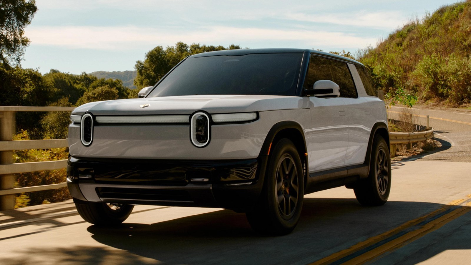 White Rivian R2 driving across a bridge on a sunny canyon road