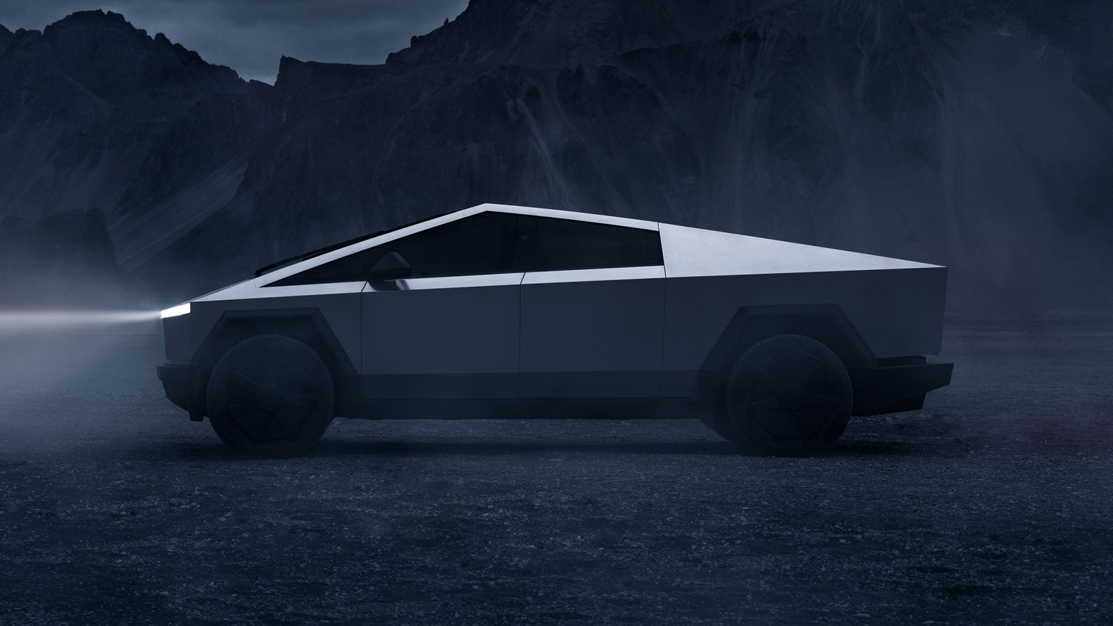 Tesla Cybertruck in profile with headlights on, parked on dark gravel below mountains