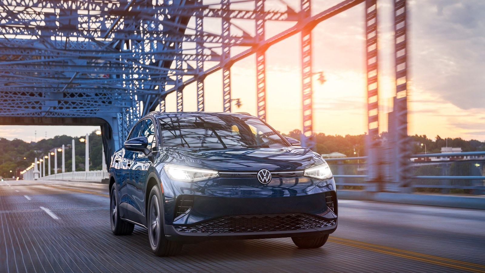Dark blue 2023 Volkswagen ID.4 driving across a steel truss bridge at sunset