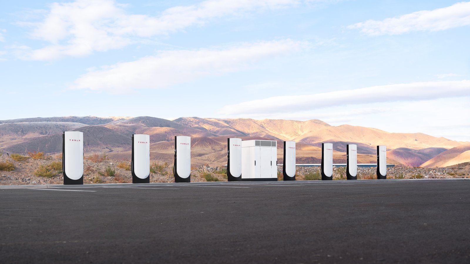 A row of white Tesla V4 Supercharger stalls with a power cabinet in an empty desert parking lot