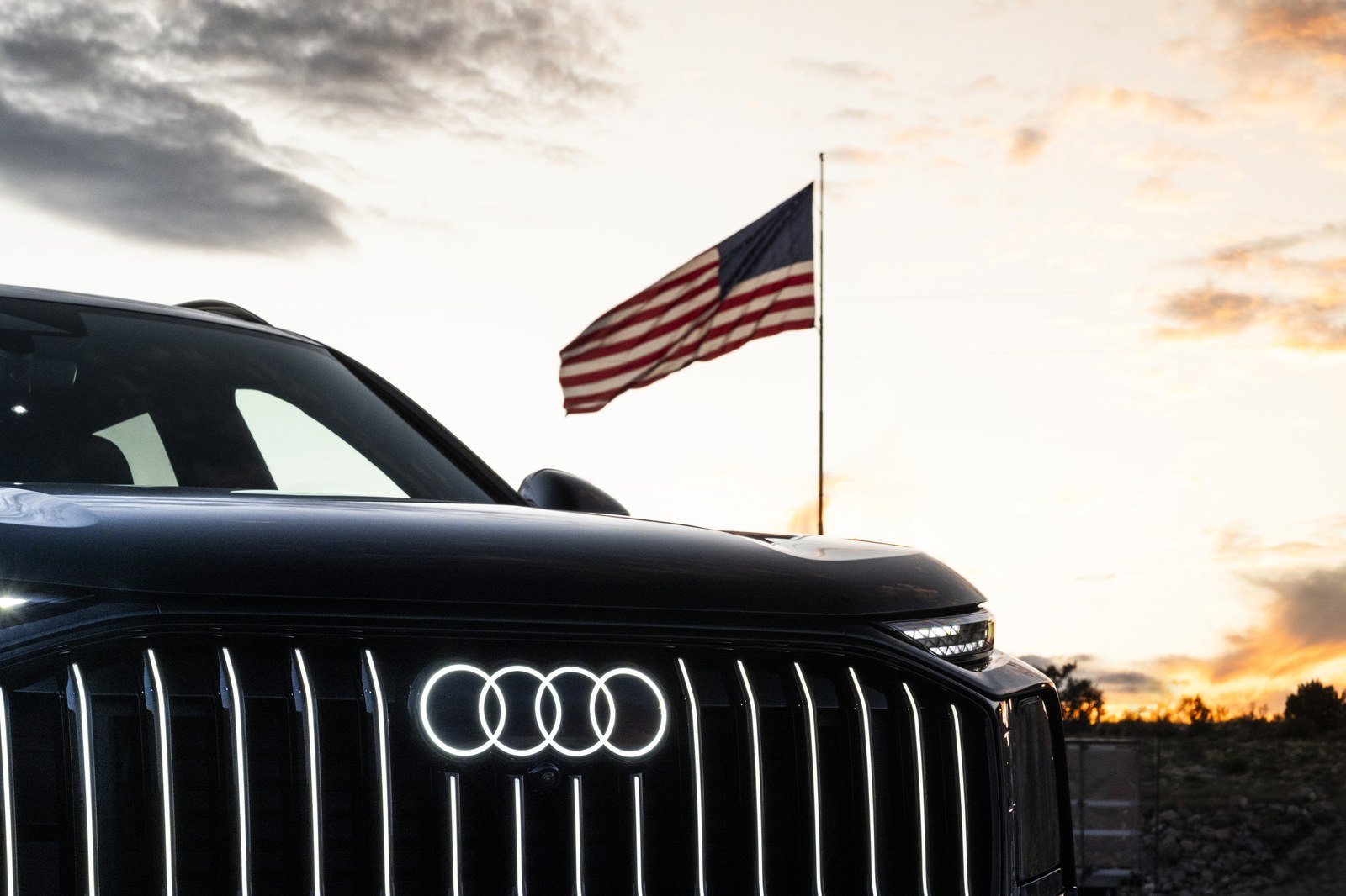 Illuminated Audi rings on the grille of a dark Audi SUV with an American flag flying behind it at sunset