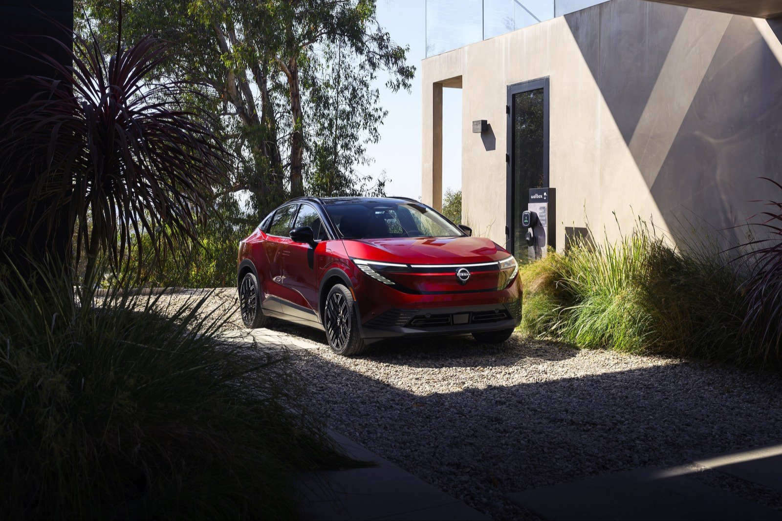A red 2026 Nissan Leaf parked on a gravel driveway next to a wall-mounted home charger