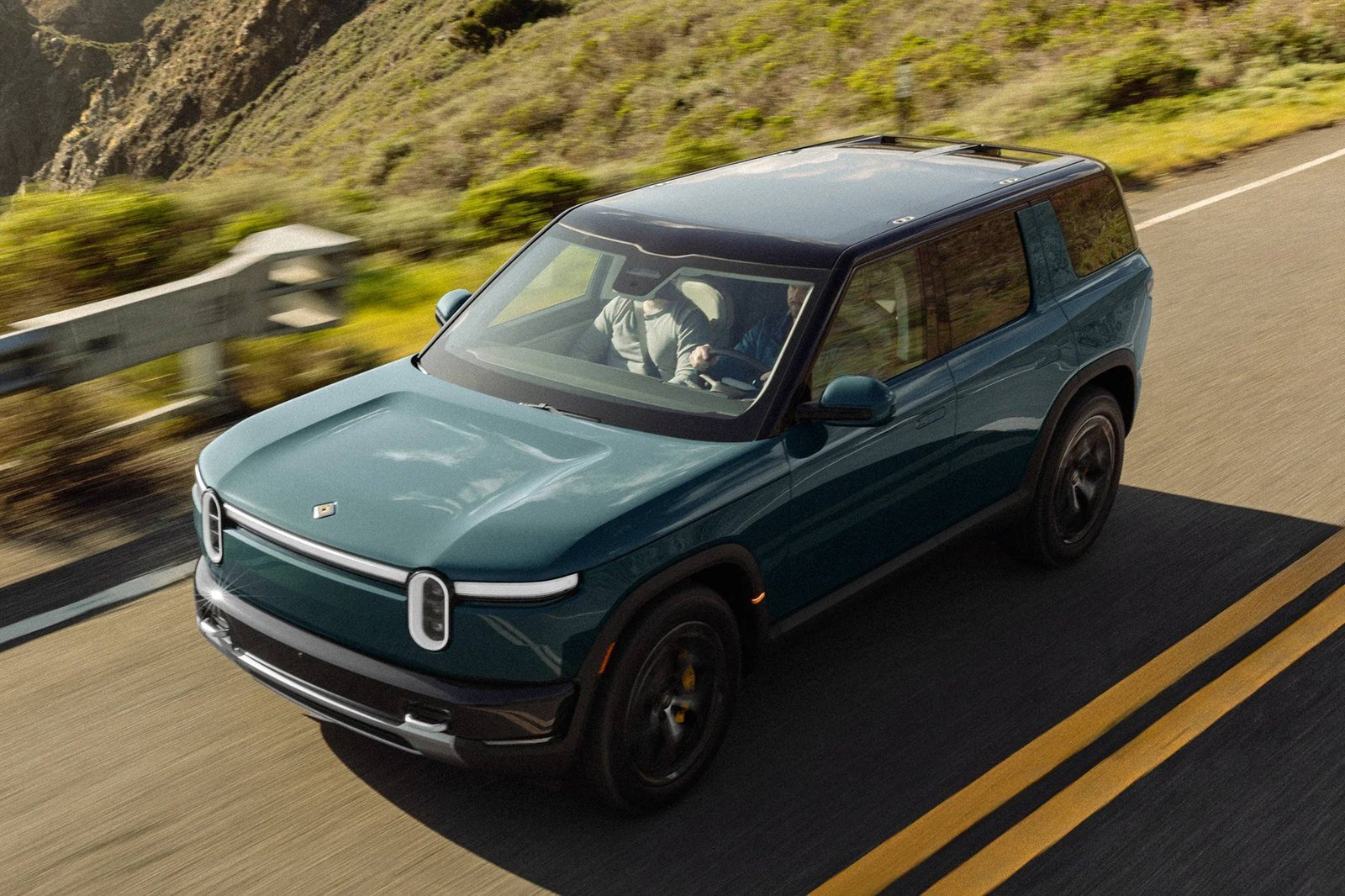 Green Rivian R2 driving on a coastal highway