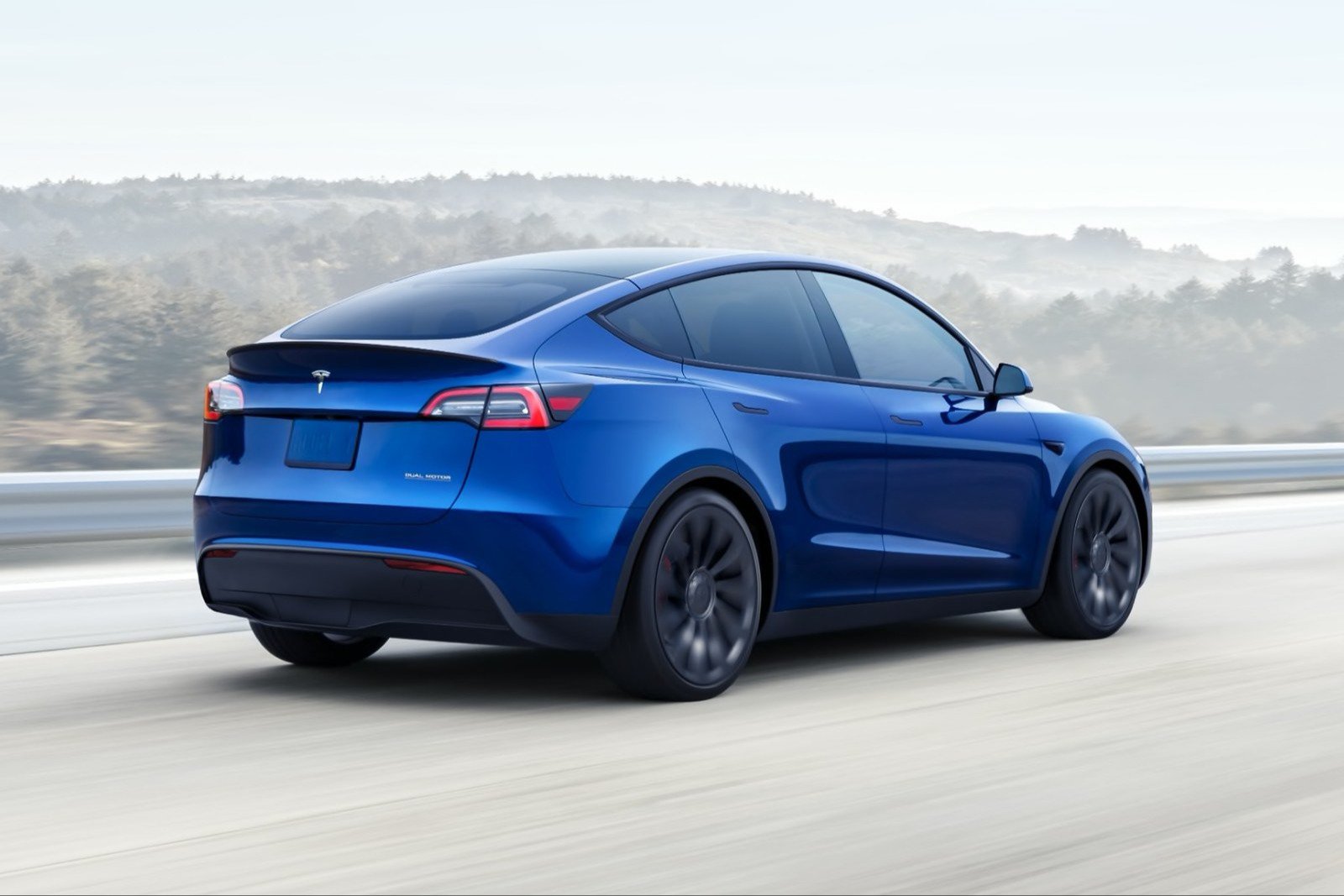 A blue Tesla Model Y driving on a highway, seen from the rear three-quarter view